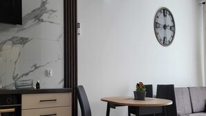 A modern kitchen with a marble backsplash, a built-in oven, and a dining area with a round table and four chairs. A wall-mounted clock is visible above the dining table. - Comfortable Holiday Apartment Close to the sea (Mielno)