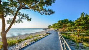 Een houten looppad langs een strand, met een hekwerk, omgeven door bomen en begroeiing.