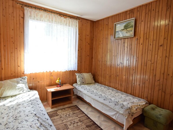 A room with wooden paneling, two single beds with floral bedding, a small wooden nightstand with a lamp, a green footstool, and a window with sheer curtains.