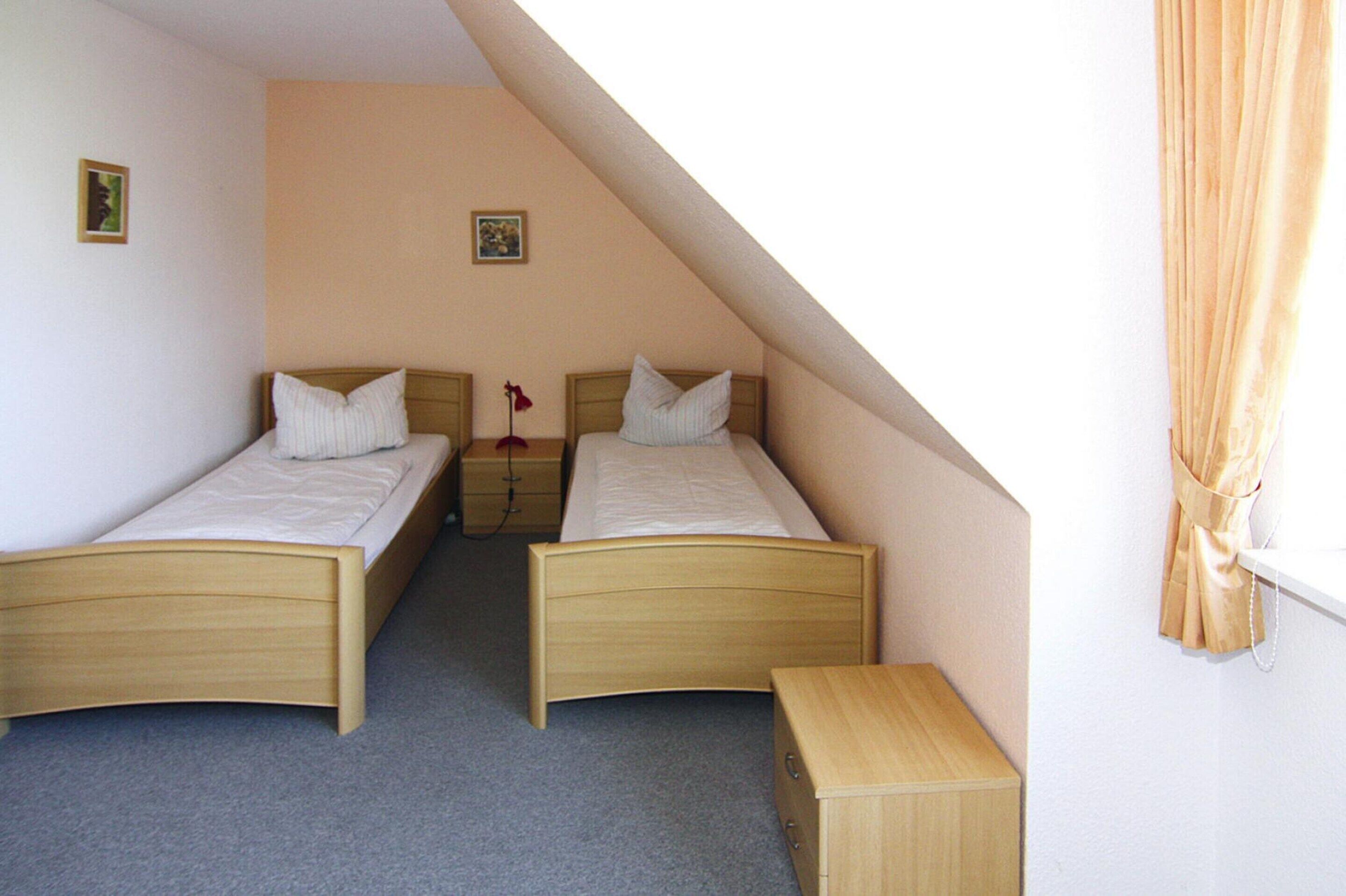A small, single-room bedroom with two single beds, a nightstand, a dresser, and a small window with curtains.