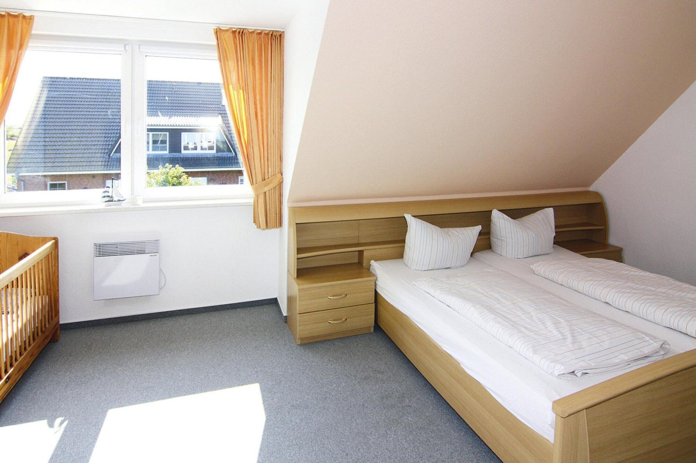 Ein Schlafzimmer mit einem hölzernen Bett, zwei Kissen, einem Nachttisch, einem Kinderbett und einem Fenster mit Vorhängen.