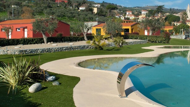 A pool with a curved edge, surrounded by a well-maintained lawn and a stone wall. In the background, there are colorful buildings and a hill with trees. - Village Mobile Home in Rebi (Imperia)