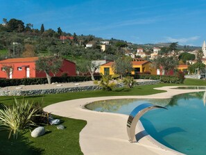 Ein Pool mit gewölbter Kante, umgeben von einem gepflegten Rasen und einer Steinsäule. Im Hintergrund sind bunte Gebäude und ein Hügel mit Bäumen zu sehen.