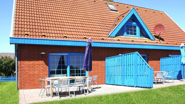 A red brick building with a blue roof and a triangular window. There is a patio with a table and chairs, and a blue fence.