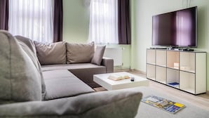 A modern living room with a sofa, a coffee table with books and a remote, a TV on a stand, and a window with curtains. - Ferienwohnung Schlossblick, Wernigerode (Wernigerode)