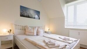 A bedroom with a large bed, white bedding, two bedside tables with lamps, a window with blinds, and a painting on the wall.