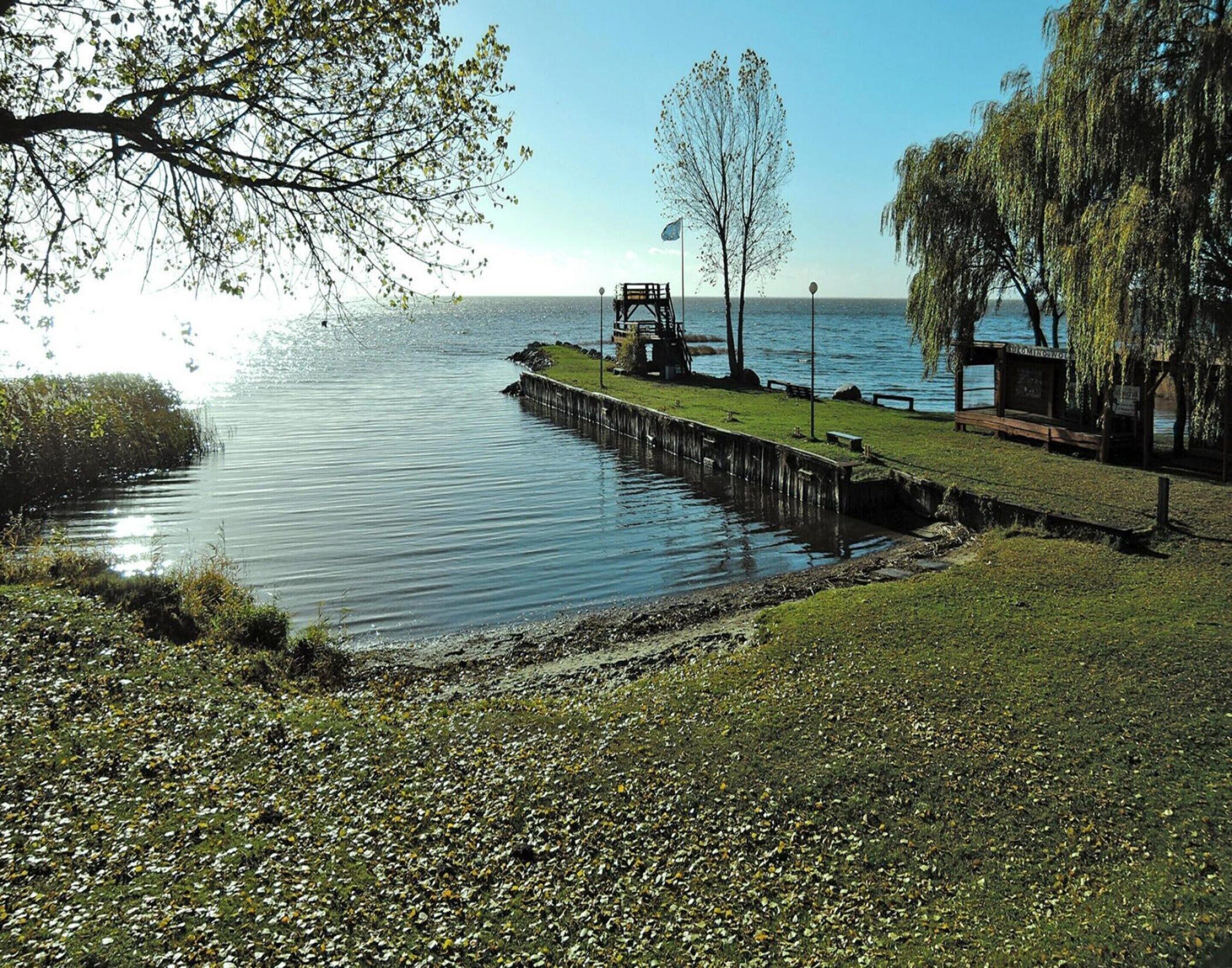 A riverside walkway with a wooden pier extending into the water, surrounded by greenery and trees.