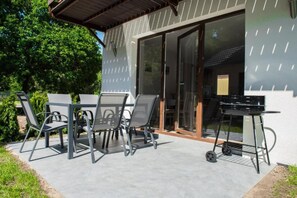 En udendørs terrasse med et bord, stole, grill og en skydedør ind til et rum indendørs.