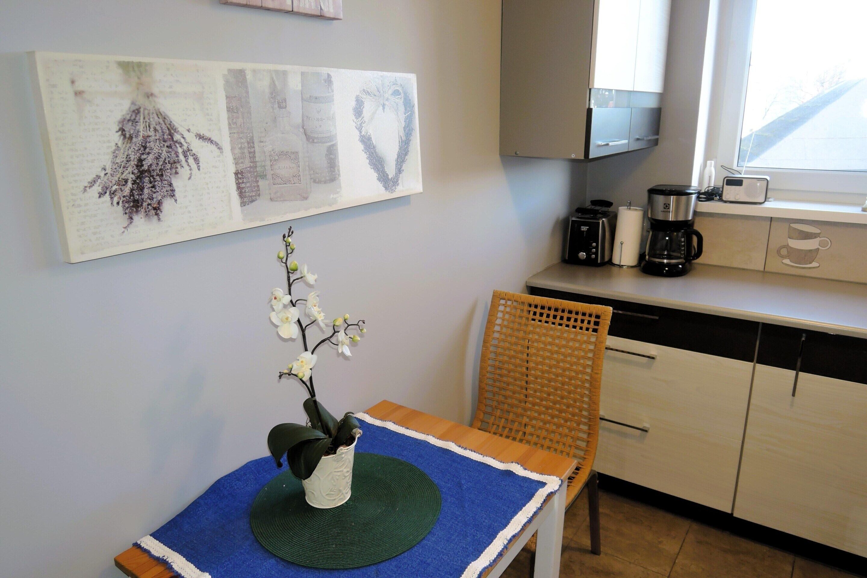 A kitchen with a coffee maker, toaster, and cabinets. There is a chair and a table with a vase of flowers.