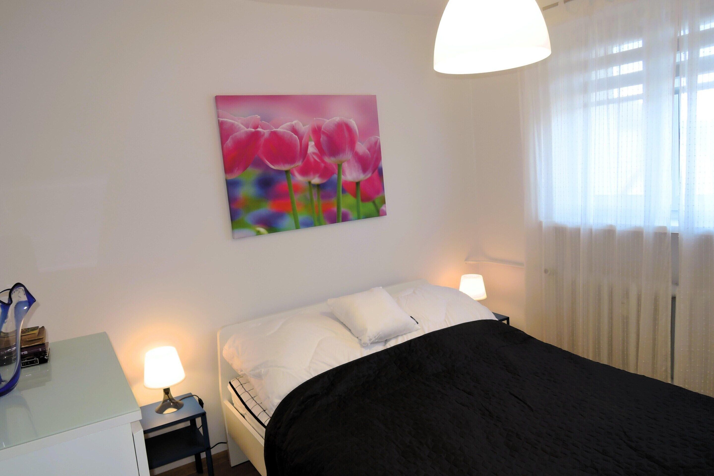A bedroom with a bed, two bedside lamps, a nightstand, a painting, and a window with curtains.