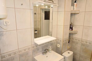 Un bagno con lavandino bianco, uno specchio e un water. Si notano mensole con articoli da toilette e un portasciugamani.