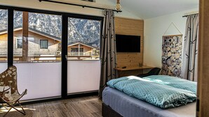 Une chambre à coucher comprenant un lit, une chaise, un bureau, une télévision et une vue sur un chalet de montagne, visible à travers de grandes fenêtres.