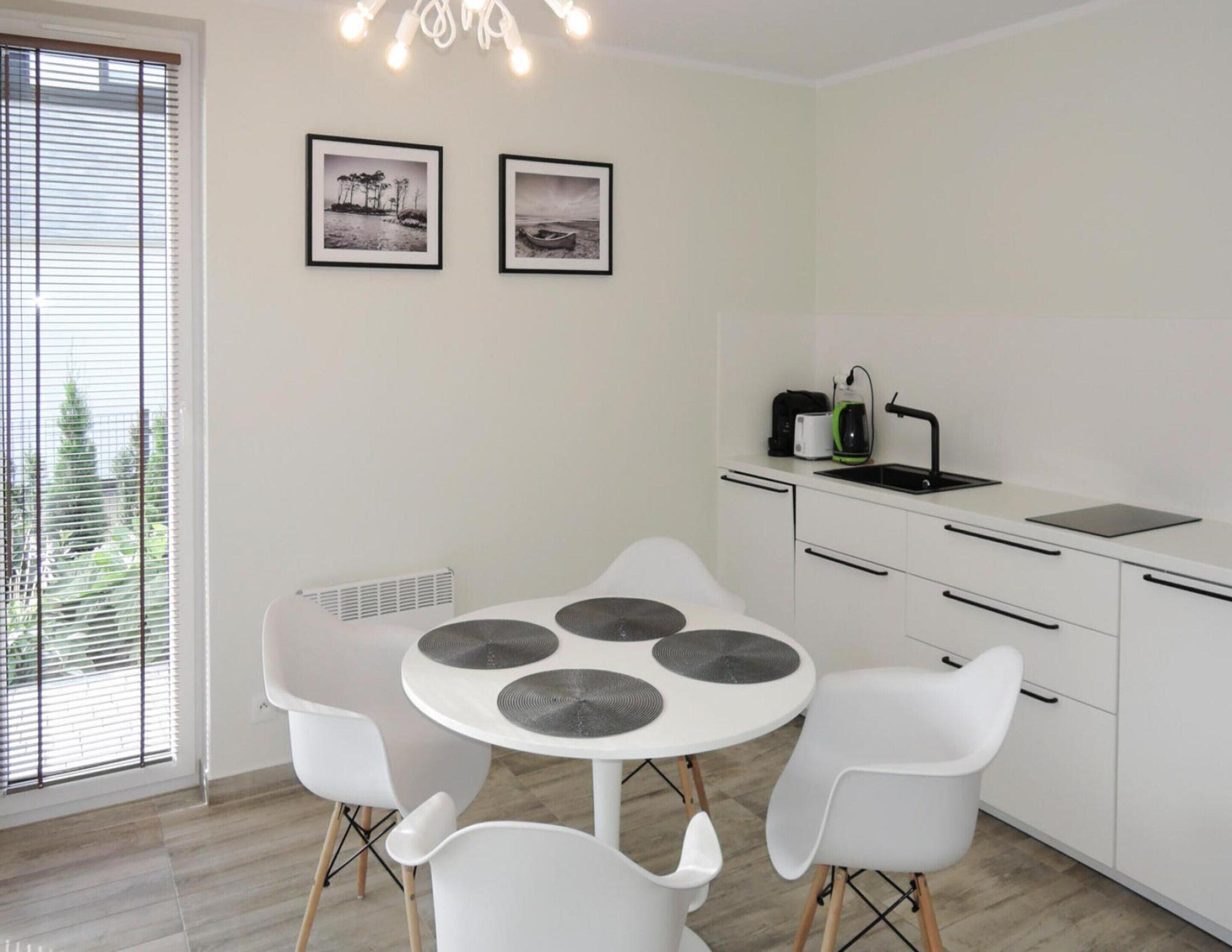 Una cocina moderna con una mesa redonda para comer, sillas blancas y un lavamanos empotrado. Hay dos cuadros en la pared y una ventana con persianas.
