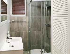 A modern bathroom with a glass-enclosed shower, a white sink, and a window with wooden blinds. - Luxury Appartment in Lukecin 60qm (Dziwnów)