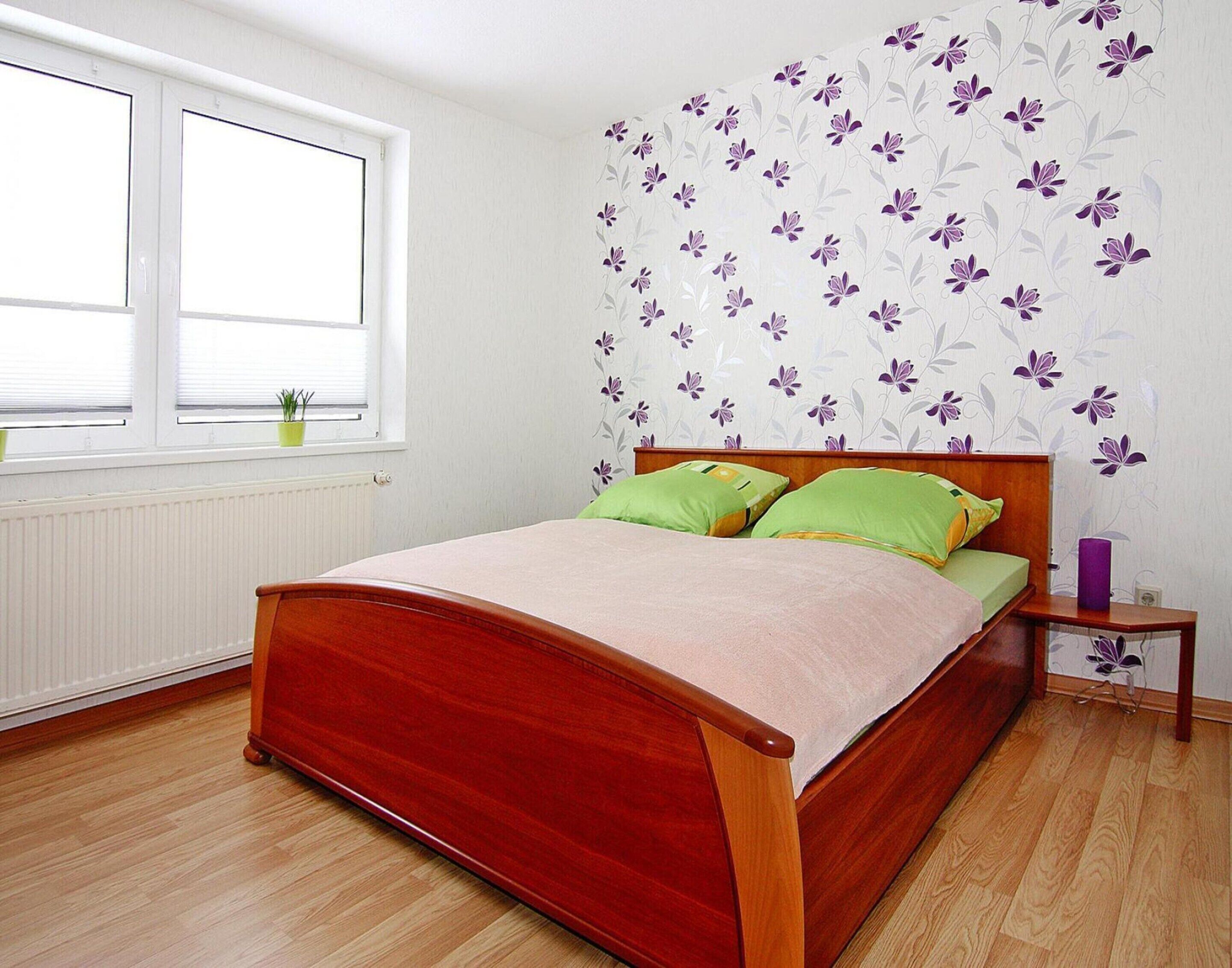 Ein Schlafzimmer mit einem hölzernen Bett, grünen Kissen, einem Nachttisch, einer violetten Vase und floralem Wandpapier.