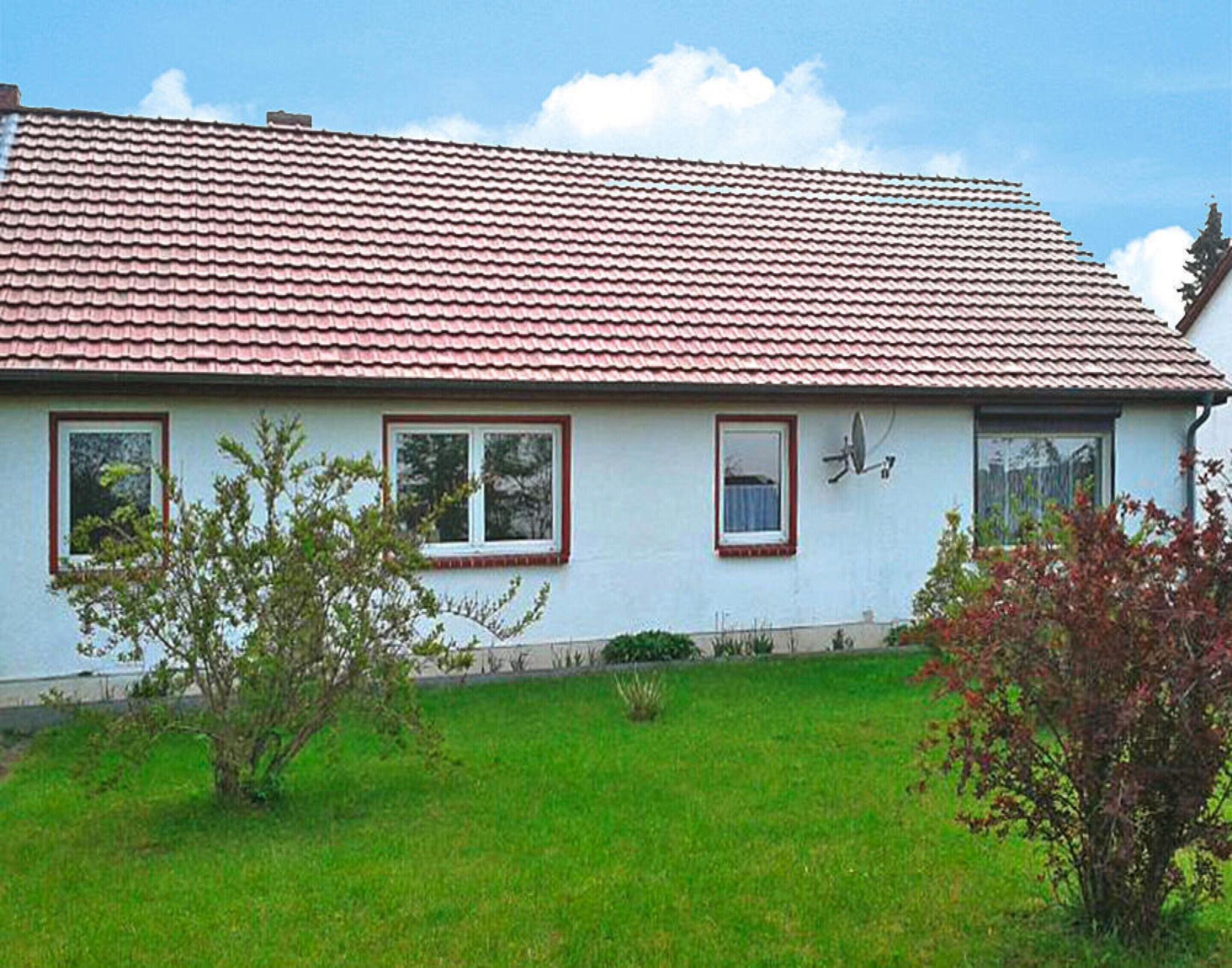 Une maison de plain-pied avec un toit recouvert de tuiles rouges, des murs extérieurs blancs et un petit jardin devant la maison, avec de la pelouse et des arbustes.