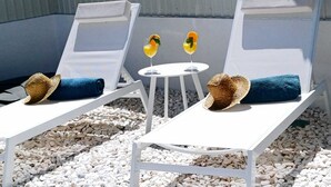 Twee witte lounge fauteuils met blauwe kussens en strooihutten, een klein rond tafeltje met twee drankjes, en een grindpad.
