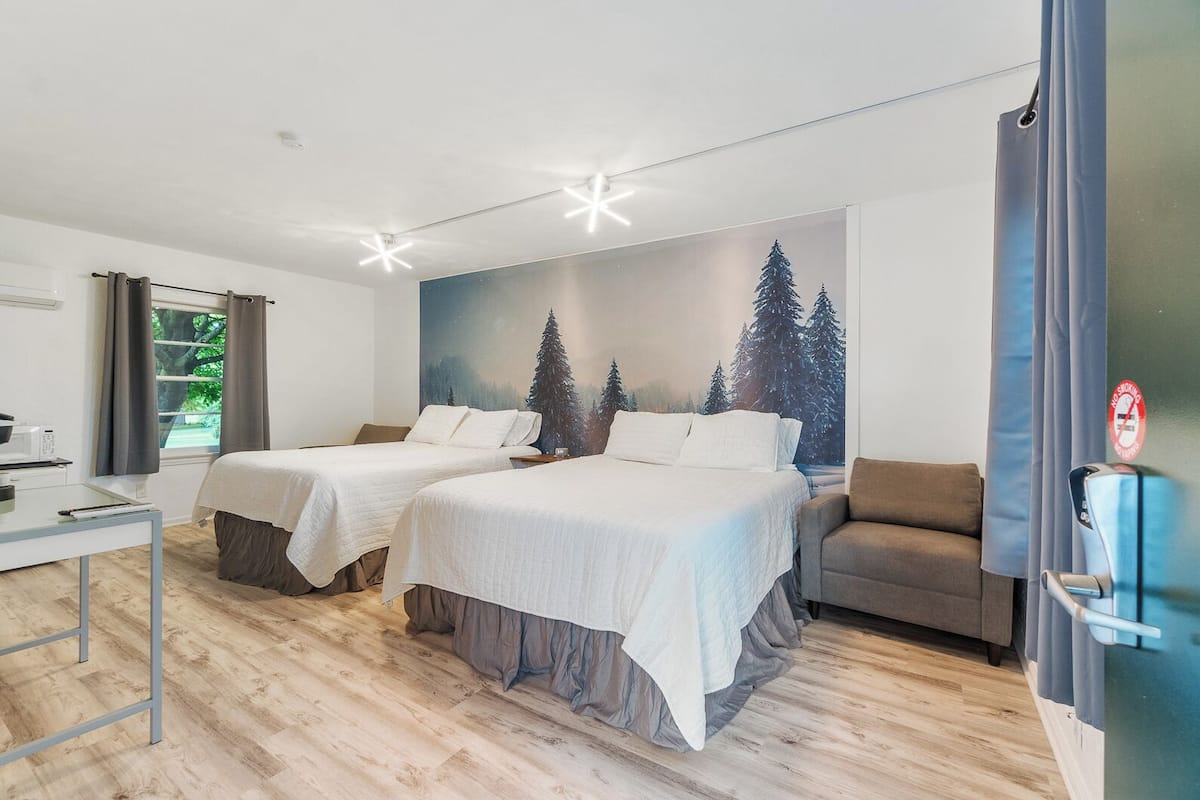 A hotel room with two beds, a desk, a chair, and a mural of trees.