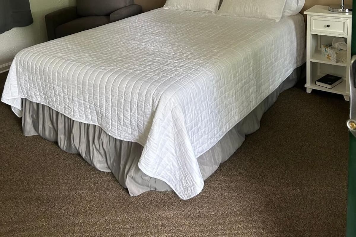 A neatly made bed with a white quilt, a bedside table with a lamp, and a chair.
