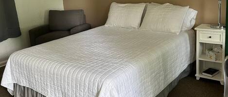 A neatly made bed with a white quilt, a bedside table with a lamp, and a chair.