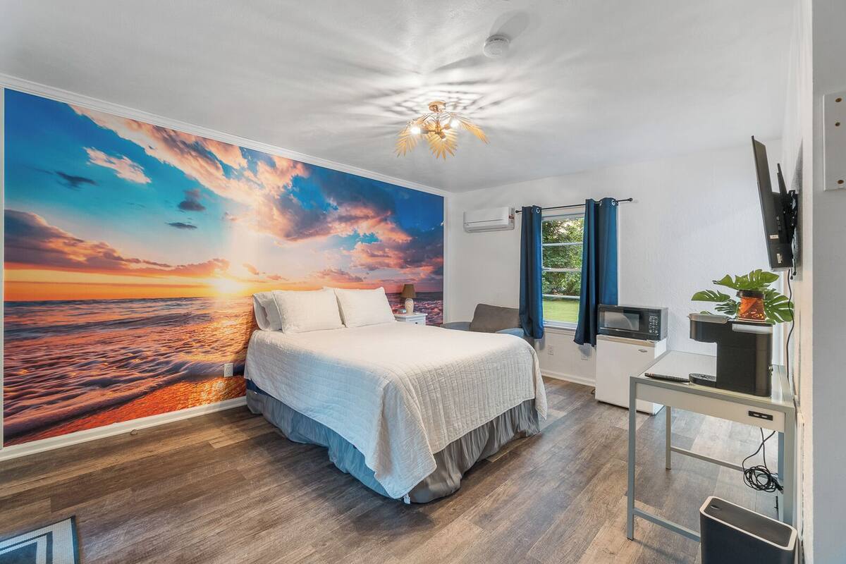 A bedroom with a large wall mural of a sunset, a bed with a white comforter, a desk with a computer, and a small plant on a table.