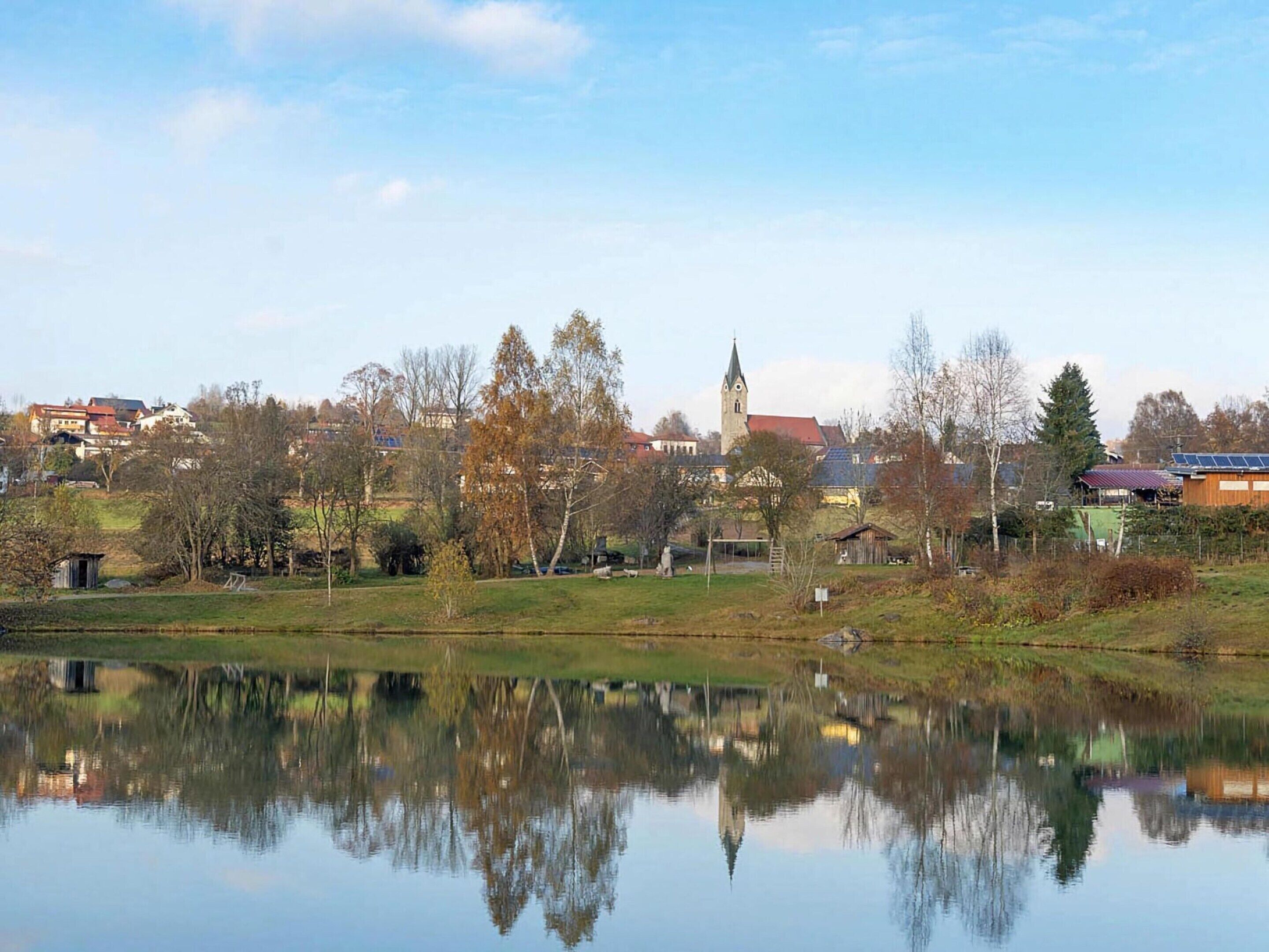 Une vue paisible sur un lac, avec un clocher d’église, des maisons et un bâtiment équipé de panneaux solaires.