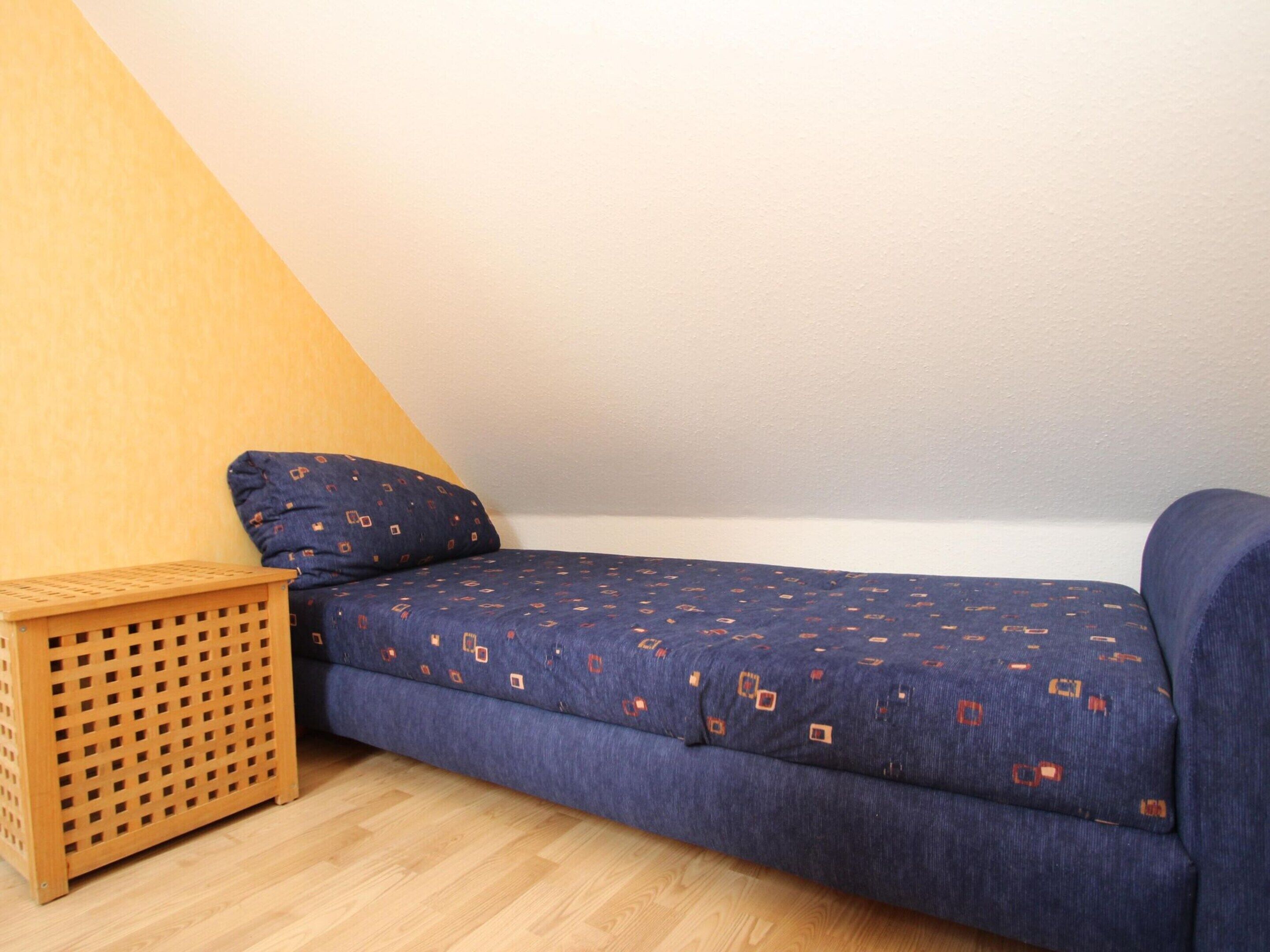 Een kleine kamer met een enkel bed, een houten vloer, een blauw bed met patroon en een houten kist.