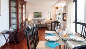Un área de comedor con una mesa de vidrio dispuesta para dos personas, un mueble de madera con puertas de cristal, un sofá y un cuadro en la pared.