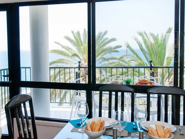 A table with a view of the sea and palm trees, two chairs, and a glass of wine.