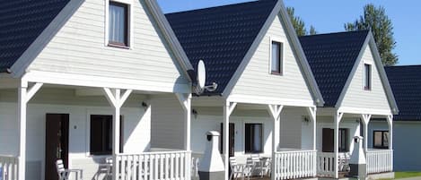 Une rangée de cabanes blanches aux toits sombres, chacune dotée d’une véranda avec des balustrades blanches et d’un petit ensemble de table et de chaises.