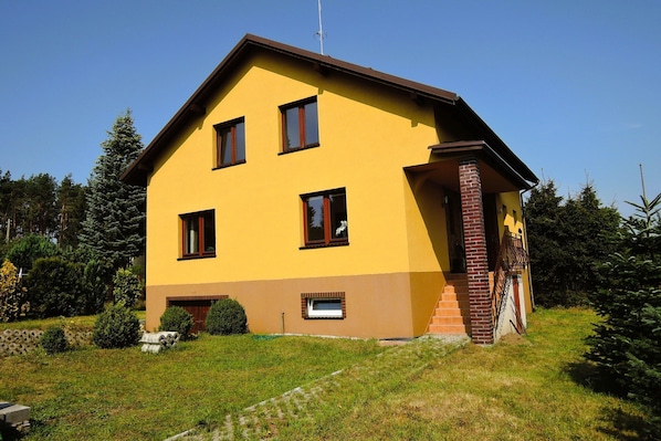 A two-story yellow building with a brick entrance, surrounded by greenery.