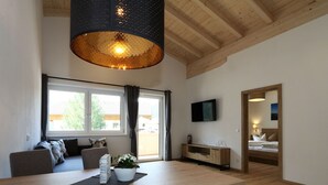A modern living room with a wooden dining table, a sofa, a TV mounted on the wall, and a large window with curtains. - Panorama Apartment Stubai (Neustift Im Stubaital)