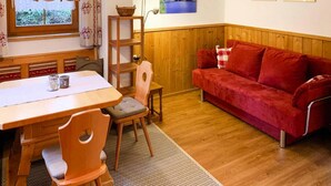 Une pièce chaleureuse avec une table à manger en bois, deux chaises, un canapé rouge et un banc en bois. Des tableaux encadrés ornent le mur, et une fenêtre est agrémentée de rideaux.