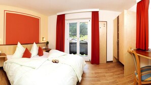 A hotel room with a large bed, a wooden desk, and a view of a forest through a sliding glass door. - Apartment in Ischgl With Parking Space (Ischgl)