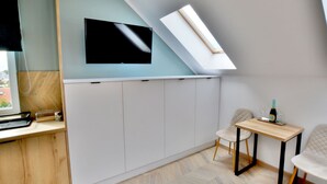 A modern room with a sloped ceiling, a flat-screen TV mounted on the wall, a wooden desk, a small table with a bottle and glasses, and a chair. - Comfortable Holiday Studio Close to the Beach (Ustronie Morskie)