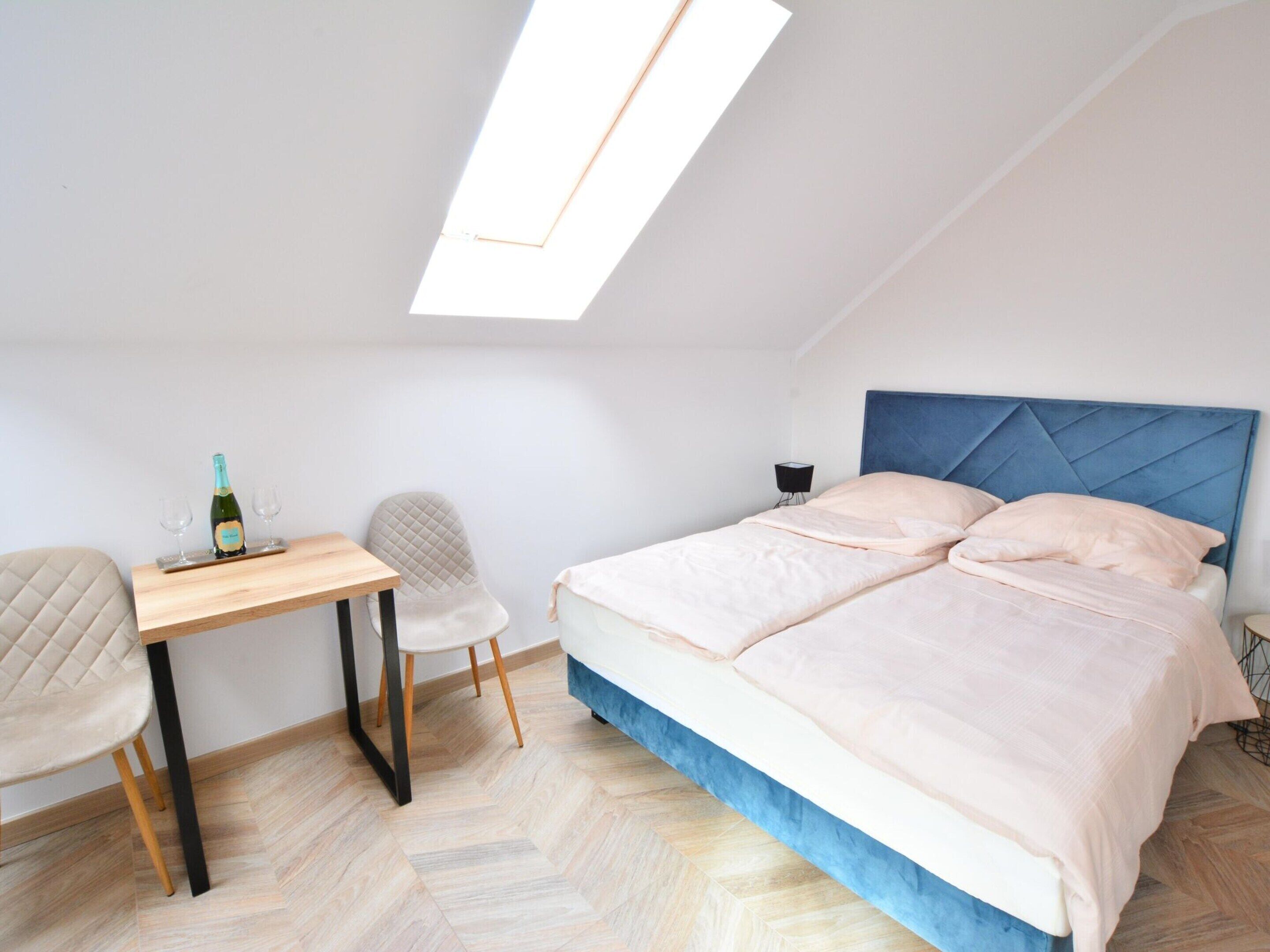 A bedroom with a bed, a desk, a chair, and a small table with a bottle and glasses.