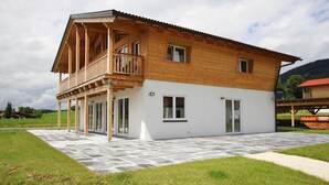 A two-story wooden house with a balcony, large windows, and a paved patio. - Ferienwohnung mit Sauna (Inzell)