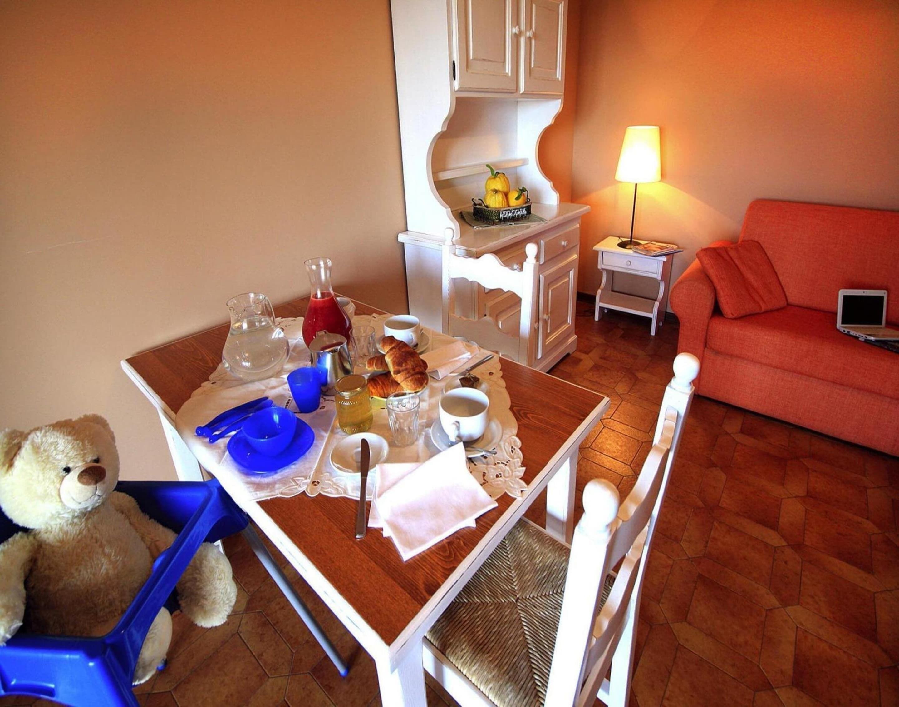 Un coin repas avec une table mise pour le petit-déjeuner, un ours en peluche et un ordinateur portable posé sur un canapé.