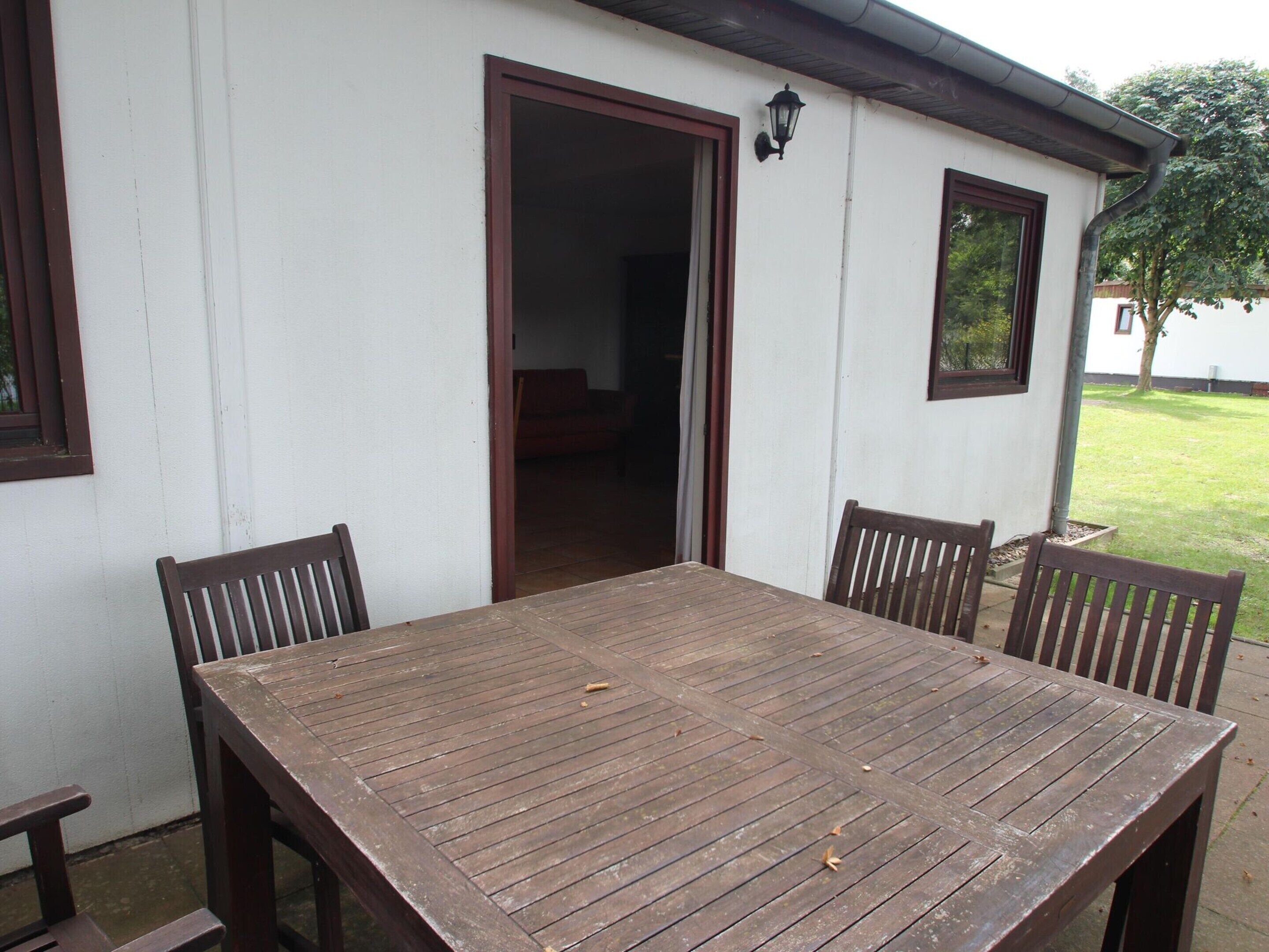 A wooden table with four chairs in front of a white building with a brown door and windows.