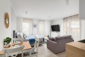 En moderne og lys stue med en sofa, et spisebord med stole, et fjernsyn og et stort vindue med lette gardiner.