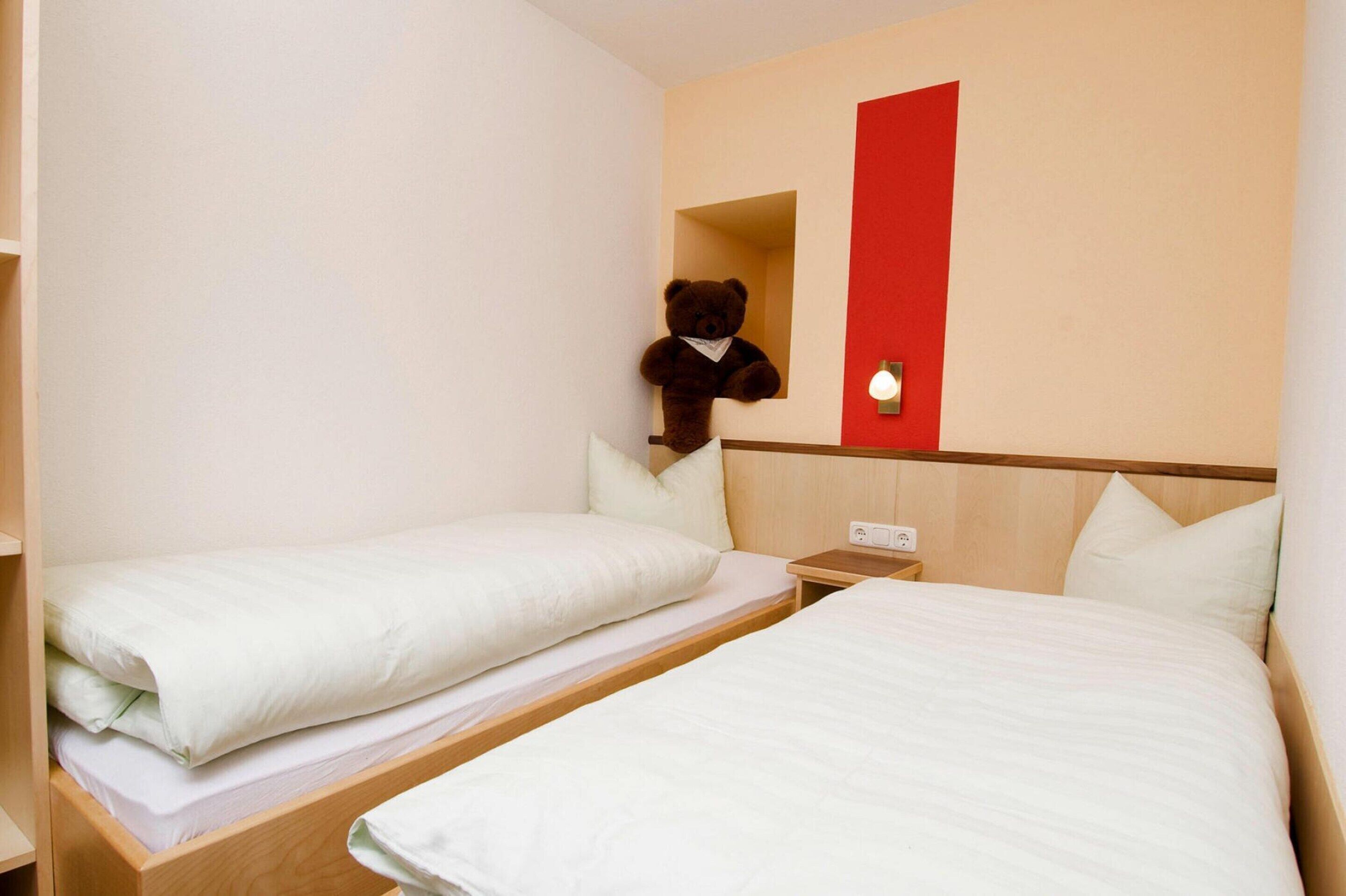 Ein Hotelzimmer mit zwei Einzelbetten, einem Holzkopfende, einer roten Akzentwand und einem Teddybär an der Wand.