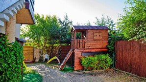 Une cabane en bois avec un toboggan et un petit escalier, entourée de verdure et d’une clôture en bois.