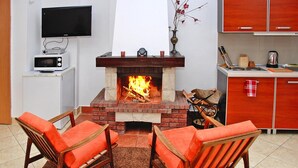 Un salon doté d’une cheminée, de deux fauteuils en bois avec des coussins orange, d’un téléviseur fixé au mur et d’un coin cuisine avec des placards et un plan de travail.