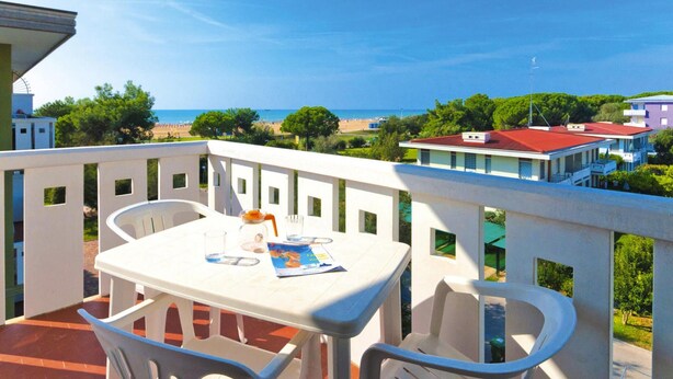 A balcony with a table and chairs, overlooking a beach and buildings. - Apartments Condominio Diana, Bibione Lido del Sole-formerly TUI Ferienhaus (San Michele al Tagliamento)