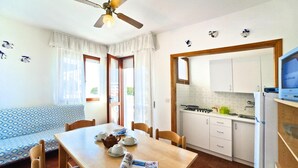A compact kitchen with white cabinets, a stove, and a refrigerator. There is a dining table with chairs and a small sofa in the living area. - Apartments Condominio Diana, Bibione Lido del Sole-formerly TUI Ferienhaus (San Michele al Tagliamento)