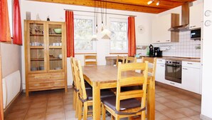 A wooden dining table with chairs, a cabinet with glass doors, a kitchen area with a stove and microwave, and a window with curtains. - Reihenhaus Dunenwaldhaus in Trassenheide (Trassenheide)
