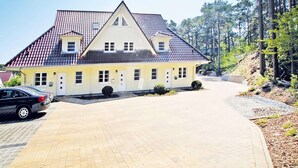 A two-story building with a tiled roof, a driveway, and a parking area with cars. - Reihenhaus Dunenwaldhaus in Trassenheide (Trassenheide)