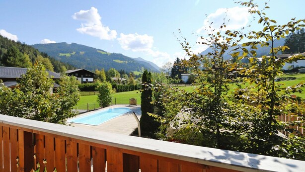 Ein Pool mit einer Holzterrasse, umgeben von Grünflächen und Häusern in einer gebirgigen Gegend.