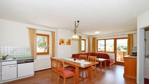 Ein Essbereich mit Holztisch und -stühlen, eine Küche mit weißen Schränken und große Fenster mit Vorhängen.