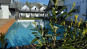 Une piscine entourée d’une terrasse en bois, elle-même bordée de bâtiments blancs dotés de balcons, sous un ciel dégagé.